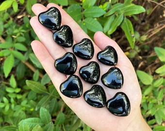 May include: A collection of twelve black obsidian heart-shaped stones. The stones are smooth and polished, and they are arranged in a hand.