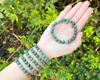 May include: A set of six stretch bracelets made of round, mottled green and brown beads. One bracelet is held in the palm of the hand, while five additional bracelets are stacked on the wrist, showcasing the natural stone texture and color.