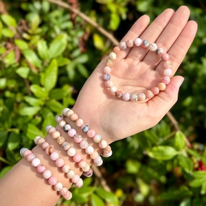 May include: A set of pink and white gemstone bracelets with round beads. The bracelets are stacked on a person's wrist.