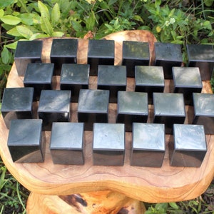 May include: A collection of 25 black obsidian cubes arranged in a grid pattern on a wooden surface. The cubes are smooth and polished, with a slight sheen.