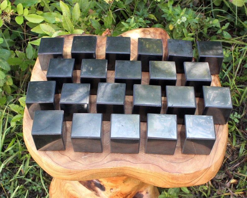 May include: A collection of 25 black obsidian cubes arranged in a grid pattern on a wooden surface. The cubes are smooth and polished, with a slight sheen.