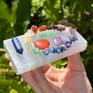 May include: A selenite charging plate with a rainbow of crystals arranged in a circle. The crystals include red jasper, carnelian, yellow calcite, green aventurine, blue sodalite, amethyst, and tiger's eye.
