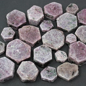 May include: A collection of raw, hexagonal ruby crystals in various sizes. These natural gemstones feature a matte, textured surface with a color palette ranging from dusty pink to deep magenta, interspersed with dark grey and black mineral inclusions.