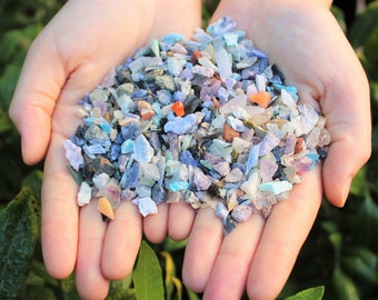 May include: A close-up of a collection of small, multi-coloured fragments, including blue, purple, green, orange, and white. The pieces are held in open hands, with a blurred green background. The fragments appear to be small, possibly recycled materials.