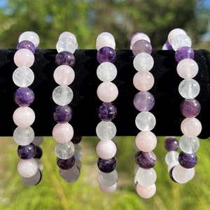 May include: Five bracelets made with amethyst, rose quartz, and clear quartz beads. The bracelets are stacked on a black surface.