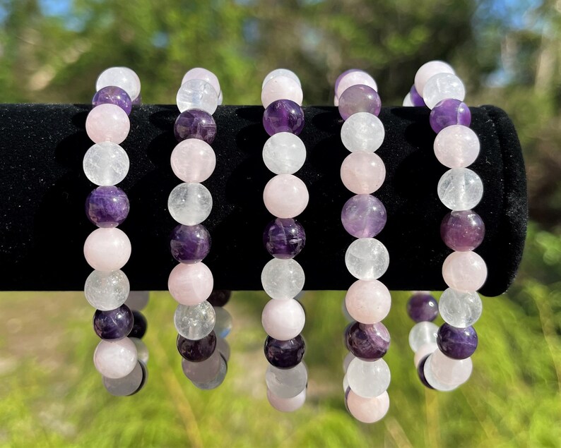 May include: Five bracelets made with amethyst, rose quartz, and clear quartz beads. The bracelets are stacked on a black surface.