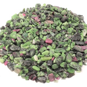 May include: A pile of green and pink gemstones, likely a mix of green zoisite and pink ruby, with some black inclusions. The stones are small and irregular in shape, and they are scattered across a white background.