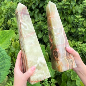 May include: Two polished stone obelisks featuring a smooth, translucent surface with banded layers of pale green, cream, and deep reddish-brown mineral inclusions. Each tower has a pointed apex and a rectangular base, showcasing natural stone patterns.