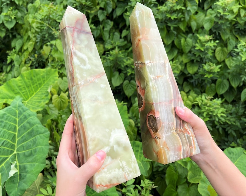 May include: Two polished stone obelisks featuring a smooth, translucent surface with banded layers of pale green, cream, and deep reddish-brown mineral inclusions. Each tower has a pointed apex and a rectangular base, showcasing natural stone patterns.
