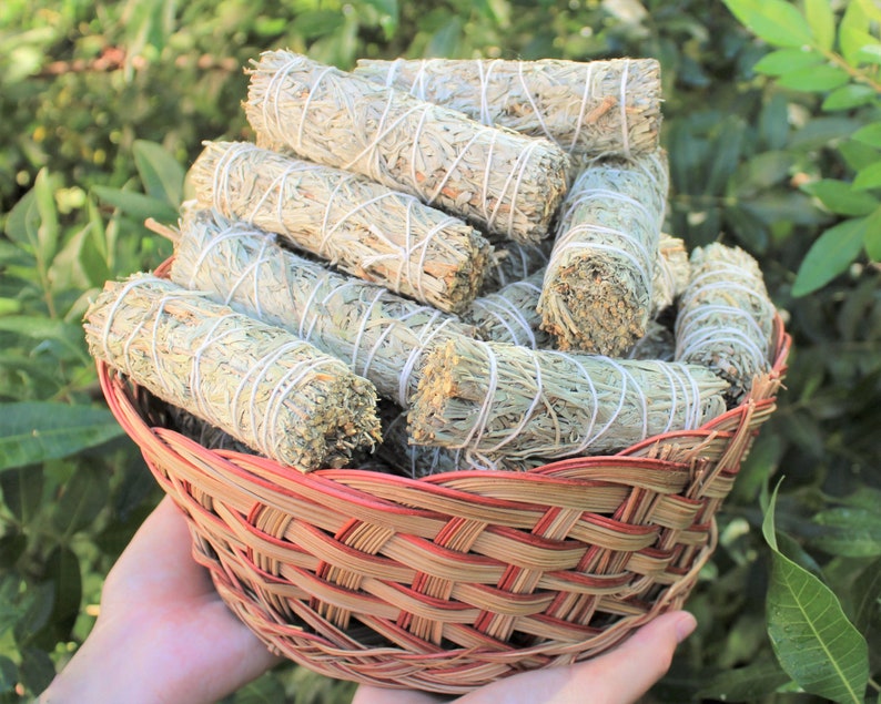 May include: A wicker basket filled with bundles of white sage smudge sticks. The sticks are tied with twine and have a natural, earthy look.