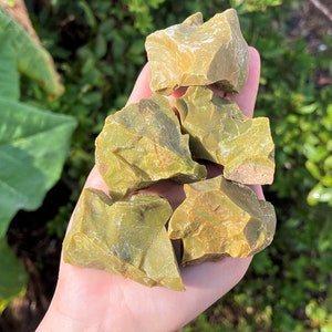 May include: A collection of five rough green opal stones. The stones are various sizes and shapes, with a translucent green color and a slight shimmer.