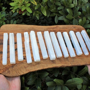 May include: A collection of 15 white selenite wands arranged in a row on a wooden board. The wands are smooth and polished, with a natural, slightly cloudy appearance.
