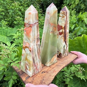 May include: Three polished onyx obelisks with natural color variations of green, orange, and brown. The obelisks are displayed on a wooden plank, showcasing their geometric shape and unique mineral patterns. The background features lush green foliage.