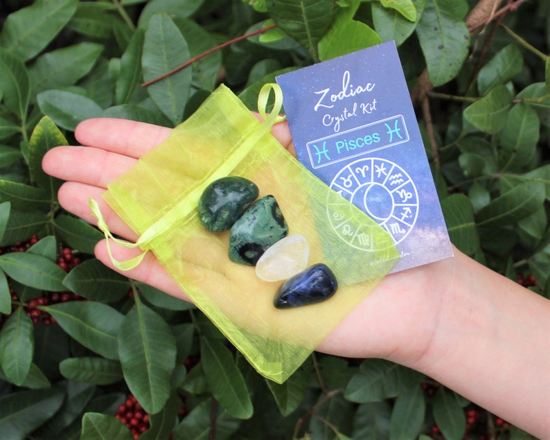 K&ouml;nnte beinhalten: Eine gelbe Organza-Tasche gef&uuml;llt mit drei Kristallen: einem gr&uuml;nen Aventurin, einem klaren Quarz und einem blauen Sodalith. Eine Karte mit dem Text "Zodiac Crystal Kit Pisces ♓" und einem Tierkreisrad ist sichtbar.