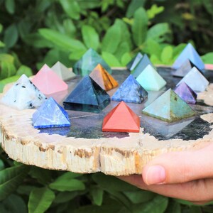 Puede incluir: Una colecci&oacute;n de peque&ntilde;as pir&aacute;mides de piedras preciosas de colores dispuestas sobre una rodaja de madera natural. Las pir&aacute;mides var&iacute;an en color, incluyendo azul, rosa, verde y rojo. La rodaja de madera tiene un borde marr&oacute;n claro y un centro gris oscuro, sostenida por manos.