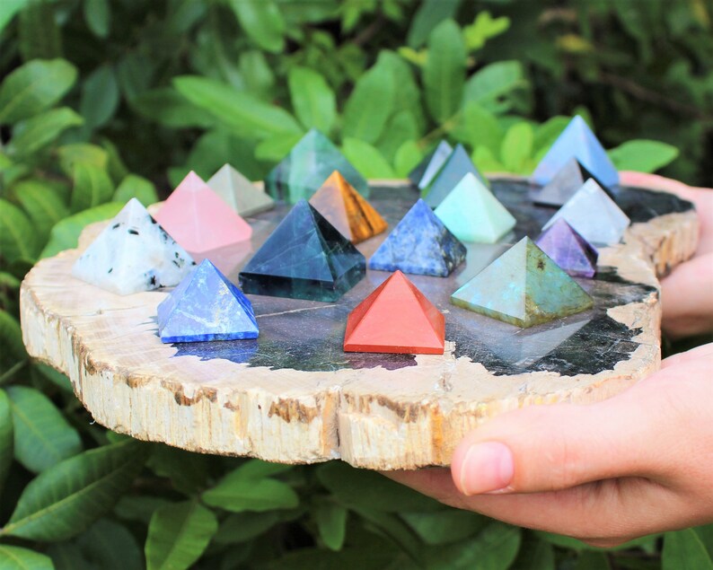 Puede incluir: Una colecci&oacute;n de peque&ntilde;as pir&aacute;mides de piedras preciosas de colores dispuestas sobre una rodaja de madera natural. Las pir&aacute;mides var&iacute;an en color, incluyendo azul, rosa, verde y rojo. La rodaja de madera tiene un borde marr&oacute;n claro y un centro gris oscuro, sostenida por manos.