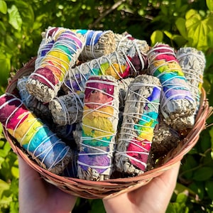 May include: A basket filled with bundles of white sage, adorned with colorful dried flower petals in a rainbow pattern. Each bundle is wrapped with white string. The basket is made of woven brown material.