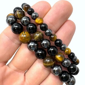 Pode incluir: Tr&ecirc;s pulseiras sobrepostas com contas pretas, olho de tigre e hematita.