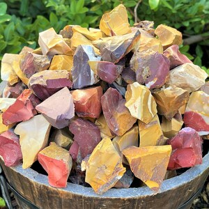 May include: A wooden bucket overflowing with a collection of raw, colorful jasper stones. The stones display a variety of colors, including red, yellow, cream, and purple. The rough-cut stones are of various sizes and shapes, creating a natural, earthy aesthetic.