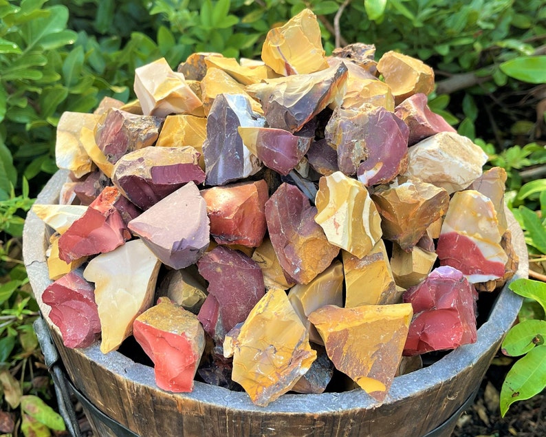 May include: A wooden bucket overflowing with a collection of raw, colorful jasper stones. The stones display a variety of colors, including red, yellow, cream, and purple. The rough-cut stones are of various sizes and shapes, creating a natural, earthy aesthetic.