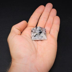 Puede incluir: Una peque&ntilde;a piedra en forma de pir&aacute;mide con un patr&oacute;n moteado de negro y gris sobre una base blanca. La piedra se sostiene en la mano abierta de una persona, mostrando su tama&ntilde;o y detalle. La pir&aacute;mide es un objeto decorativo.