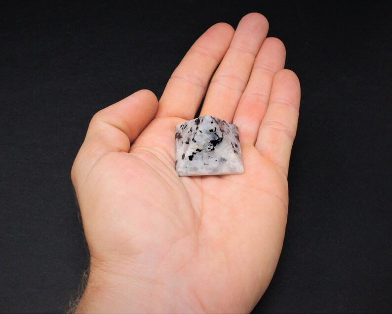 Puede incluir: Una peque&ntilde;a piedra en forma de pir&aacute;mide con un patr&oacute;n moteado de negro y gris sobre una base blanca. La piedra se sostiene en la mano abierta de una persona, mostrando su tama&ntilde;o y detalle. La pir&aacute;mide es un objeto decorativo.