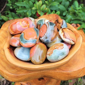 May include: A wooden bowl filled with polished stones. The stones are oval-shaped and feature a mix of colors, including orange, brown, green, and blue. The bowl is made of light-colored wood and has a natural, organic shape. The background is blurred with green foliage.
