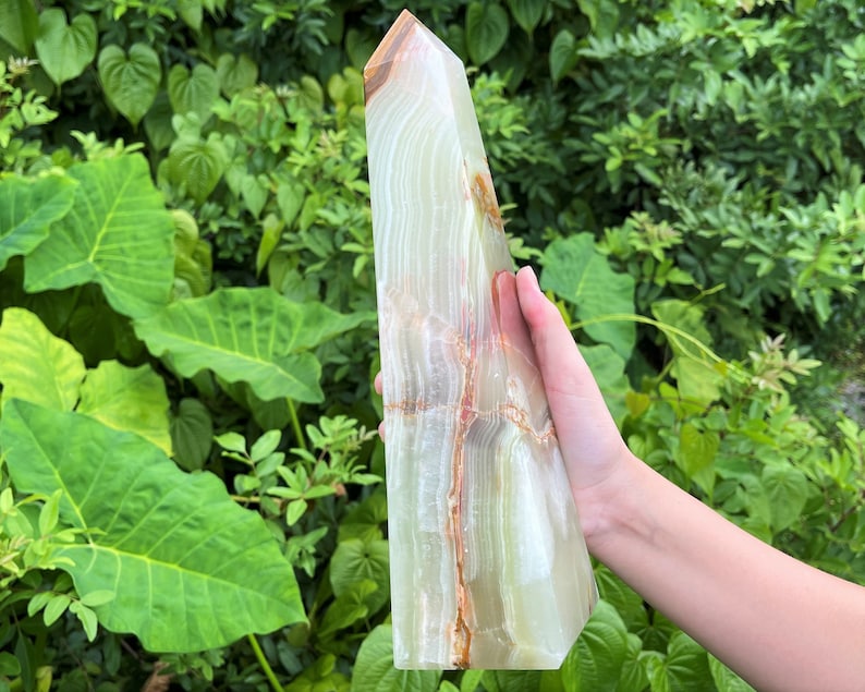 May include: A tall, polished stone tower with a hexagonal base and a pointed top. The stone features translucent layers of pale green and white, accented by prominent, jagged veins of deep orange and brown running vertically through the structure.