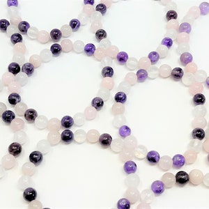 May include: A set of four bracelets made with amethyst, rose quartz, and white moonstone beads. The bracelets are stacked on a white surface.