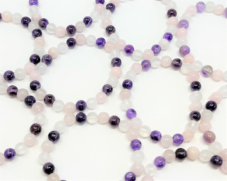 May include: A set of four bracelets made with amethyst, rose quartz, and white moonstone beads. The bracelets are stacked on a white surface.