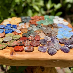 May include: A collection of polished gemstones in various colors, including blue, green, red, and purple, arranged on a wooden surface. The stones are oval and round, with a natural, earthy appearance. The arrangement suggests a display of crystals or healing stones.