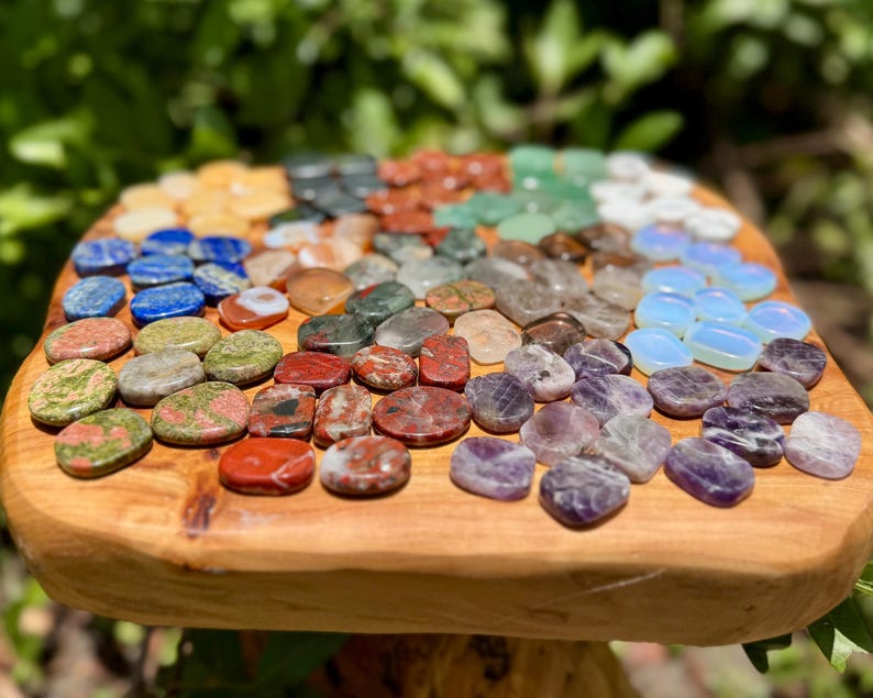 May include: A collection of polished gemstones in various colors, including blue, green, red, and purple, arranged on a wooden surface. The stones are oval and round, with a natural, earthy appearance. The arrangement suggests a display of crystals or healing stones.