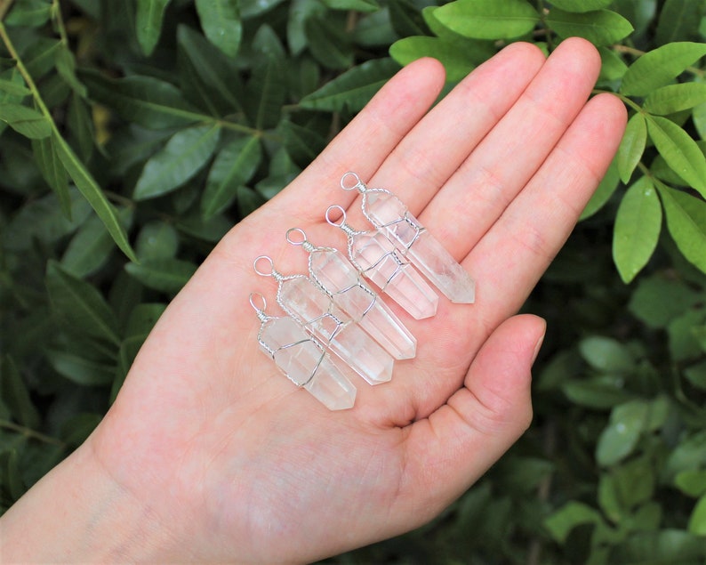 May include: Five clear quartz crystal points wrapped in silver wire with a loop at the top for hanging. The crystals are arranged in a row on a hand.