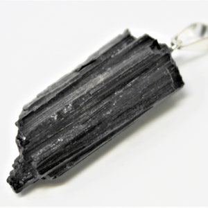 May include: A black tourmaline pendant with a silver-colored bail. The raw, natural stone has a textured surface with visible ridges and a slightly uneven shape. The pendant is set against a white background.