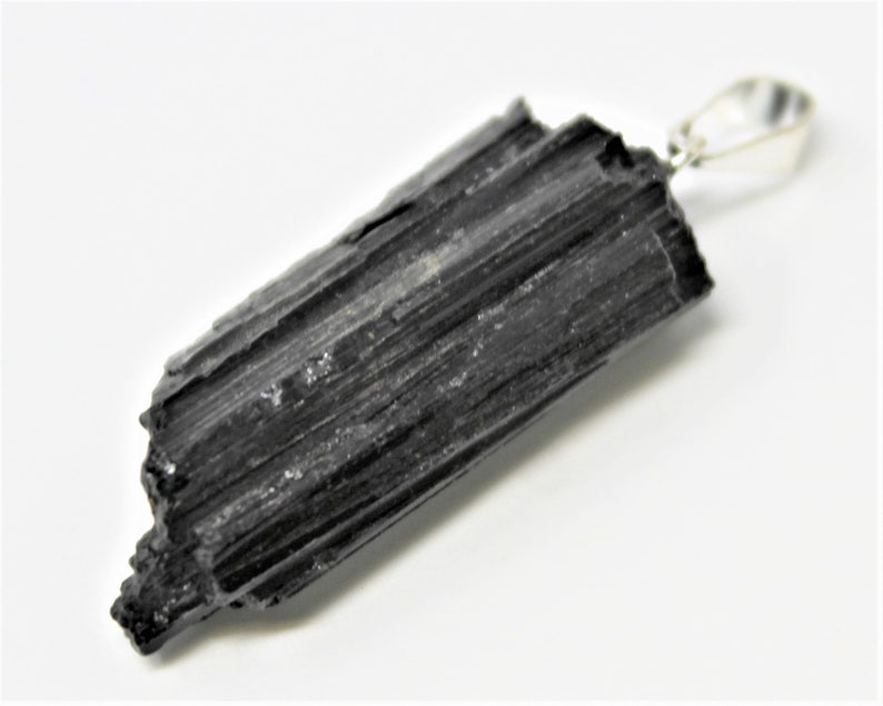 May include: A black tourmaline pendant with a silver-colored bail. The raw, natural stone has a textured surface with visible ridges and a slightly uneven shape. The pendant is set against a white background.