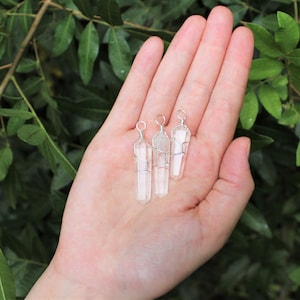 May include: Three clear quartz crystal pendants wrapped in silver wire. The pendants are hanging from a hand.