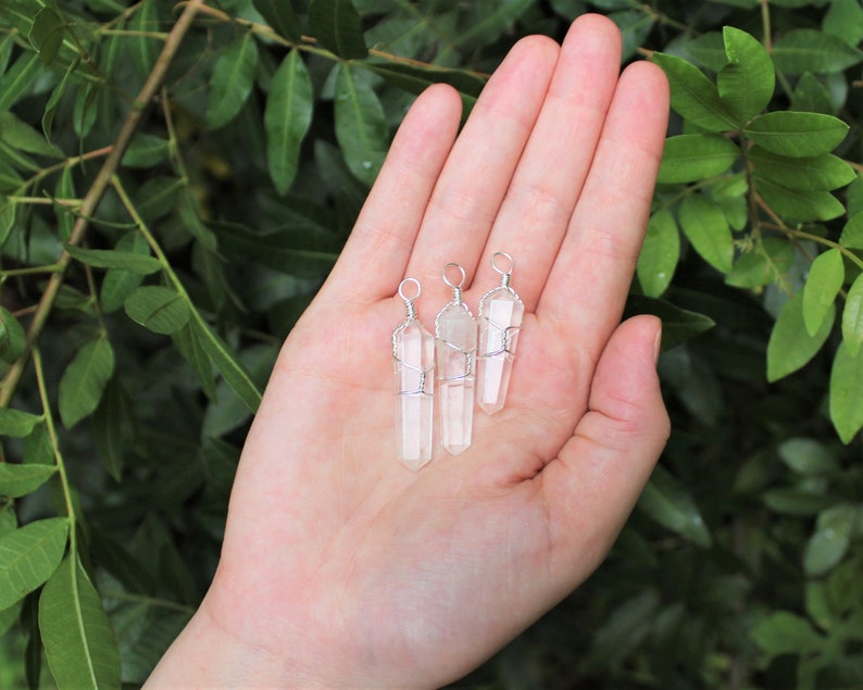 May include: Three clear quartz crystal pendants wrapped in silver wire. The pendants are hanging from a hand.
