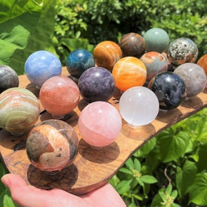 May include: A collection of 18 polished gemstone spheres displayed on a wooden tray. The spheres are various colors, including green, blue, pink, purple, orange, and brown.