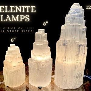 Może przedstawiać: Trzy podświetlane lampy z selenu o r&oacute;żnych rozmiarach: 15 cm, 20 cm i 30 cm wysokości. Lampy są białe i mają warstwową, krystaliczną strukturę. Tekst "SELENITE LAMPS" jest wyświetlany nad lampami.