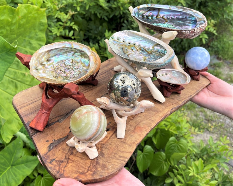 May include: A collection of three iridescent abalone shells and three polished stone spheres displayed on carved wooden stands. The items are arranged on a rustic wooden board, featuring a metallic pyrite sphere, a green banded onyx sphere, and a light blue sphere.
