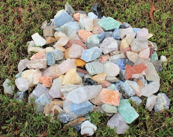 May include: A large pile of colourful, rough-cut gemstones, including clear quartz, amethyst, rose quartz, and green aventurine. The stones are scattered on a bed of green grass.