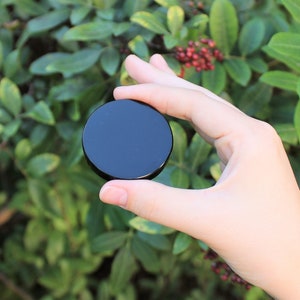 May include: A black round obsidian stone, also known as black glass, is held in a hand against a green leafy background.