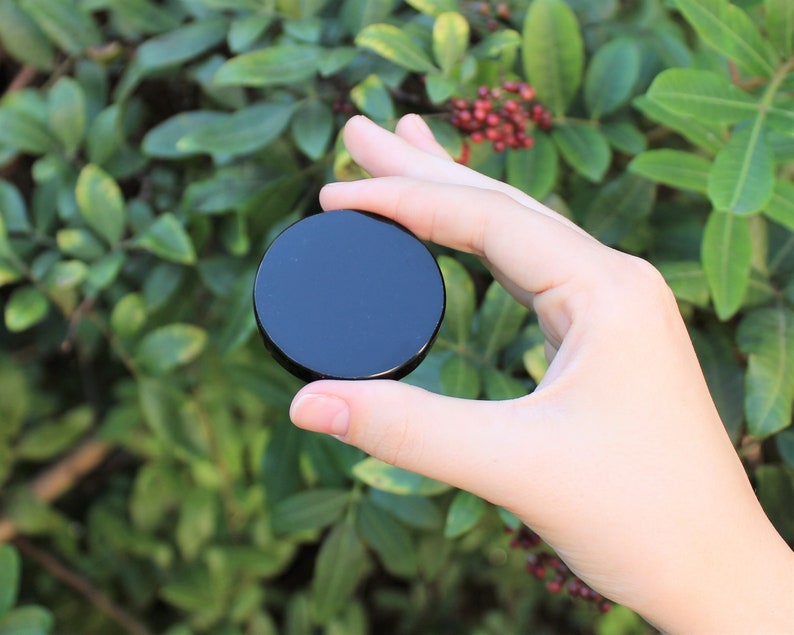 May include: A black round obsidian stone, also known as black glass, is held in a hand against a green leafy background.