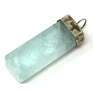 May include: A rectangular, light blue aquamarine pendant with a silver-colored cap and loop. The gemstone has a translucent quality and a slightly rough texture. The pendant is set against a white background.
