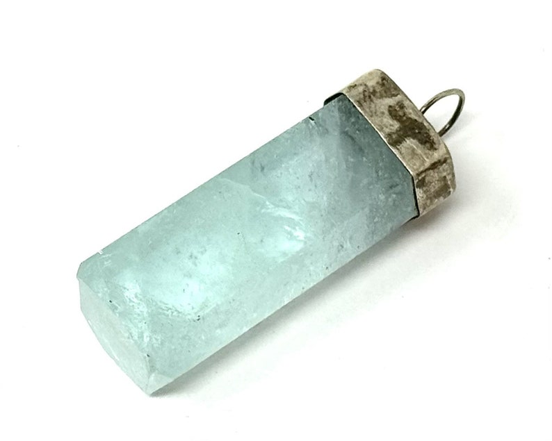 May include: A rectangular, light blue aquamarine pendant with a silver-colored cap and loop. The gemstone has a translucent quality and a slightly rough texture. The pendant is set against a white background.