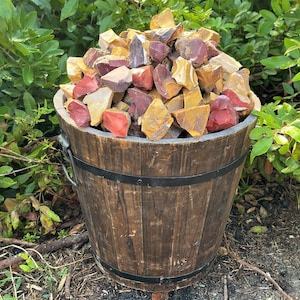 May include: A wooden bucket filled with a variety of colorful rocks. The rocks are in shades of red, yellow, and brown. The bucket is made of dark wood and has metal bands around it. The background features green foliage.
