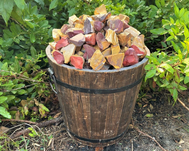 May include: A wooden bucket filled with a variety of colorful rocks. The rocks are in shades of red, yellow, and brown. The bucket is made of dark wood and has metal bands around it. The background features green foliage.