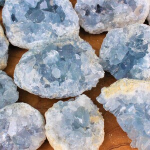 May include: A collection of blue celestite crystal clusters. The crystals are a light blue color and have a rough, natural texture. The clusters are various sizes and shapes.