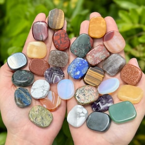 May include: A pair of hands holding a collection of polished gemstones. The stones vary in color, including red, yellow, blue, green, and brown. Some stones have unique patterns and textures, with a natural green background.
