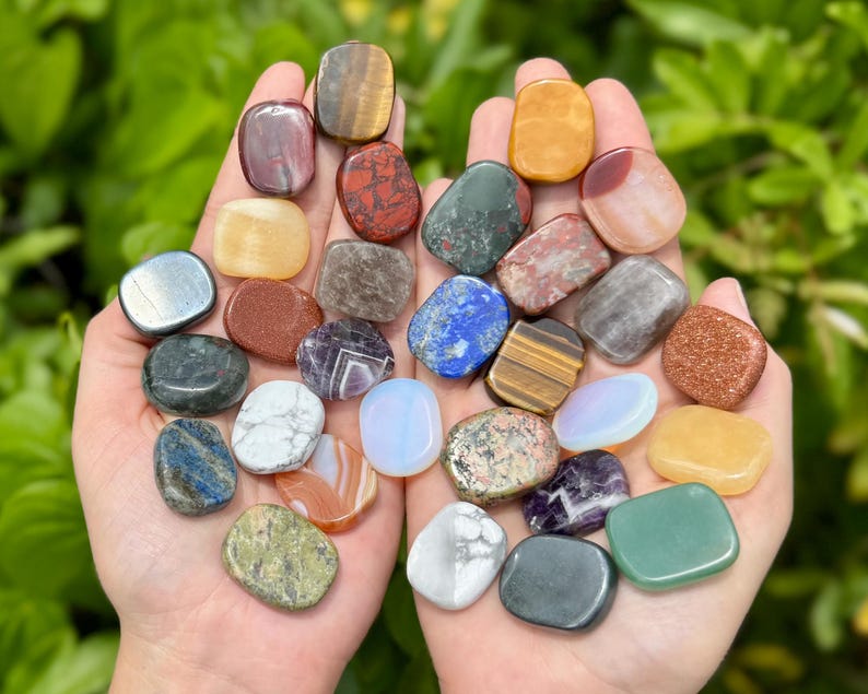 May include: A pair of hands holding a collection of polished gemstones. The stones vary in color, including red, yellow, blue, green, and brown. Some stones have unique patterns and textures, with a natural green background.